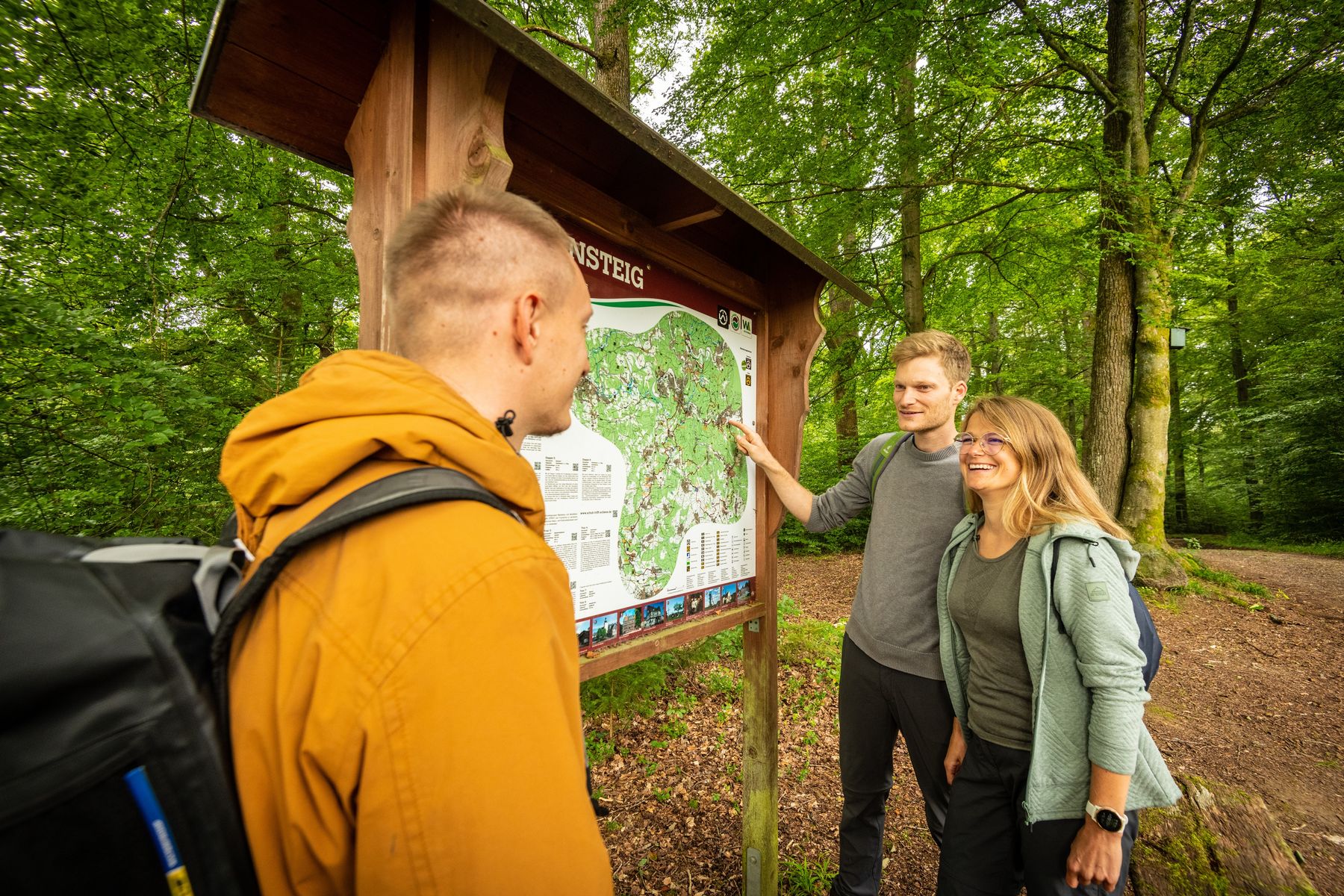 Three people with backpacks stand at a large trail map sign in a lush, green forest. One person points at the map while the others look and smile, appearing to plan their route.